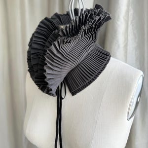 May include: A black pleated collar with a layered design, displayed on a white mannequin. The collar has a rounded shape and is secured with a black tie. The fabric appears to be a dark grey colour.