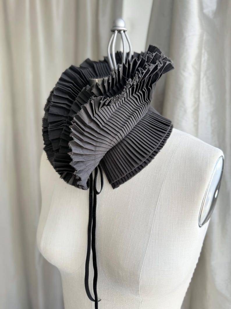 May include: A black pleated collar with a layered design, displayed on a white mannequin. The collar has a rounded shape and is secured with a black tie. The fabric appears to be a dark grey colour.