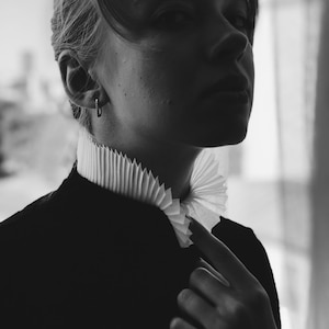 Puede incluir: Una fotografía en blanco y negro de una persona que lleva una chaqueta negra con un cuello blanco con volantes. La persona está mirando hacia un lado y tiene una expresión seria.