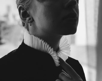 May include: A black and white photo of a person wearing a black jacket with a white ruffled collar. The person is looking to the side and has a serious expression.