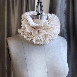 May include: A cream-coloured, ruffled collar on a dress form. The collar is made of layered fabric, creating a voluminous, textured effect. The dress form is a neutral beige colour, set against a soft grey backdrop.