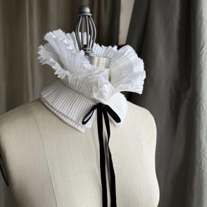 May include: A white pleated collar with a ruffled edge and a black velvet ribbon tie. The collar is displayed on a mannequin. The collar is a fashion accessory. The collar's design and texture are visible.
