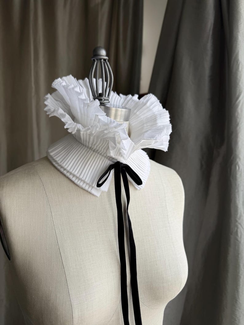 May include: A white pleated collar with a ruffled edge and a black velvet ribbon tie. The collar is displayed on a mannequin. The collar is a fashion accessory. The collar's design and texture are visible.