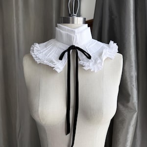 May include: A white pleated collar with a black velvet ribbon tie, displayed on a mannequin. The collar has a layered design with ruffled edges, creating a vintage or Victorian aesthetic. The black ribbon hangs down the front.
