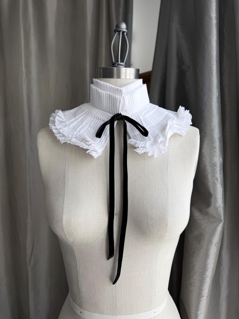 May include: A white pleated collar with a black velvet ribbon tie, displayed on a mannequin. The collar has a layered design with ruffled edges, creating a vintage or Victorian aesthetic. The black ribbon hangs down the front.