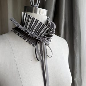 以下が含まれることがあります： 黒と白の縦縞模様が特徴のプリーツ加工されたハイネックカラー。このアクセサリーにはフリル状の縁取りがあり、中央から垂れ下がる長く細いグレーのベルベットリボンが、ネックラインでシンプルな蝶結びに結ばれています。