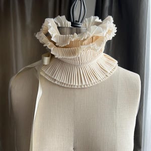 May include: A cream-coloured, pleated collar with a high neck and layered ruffles. A cream-coloured ribbon is attached to the side. The collar is displayed on a mannequin.