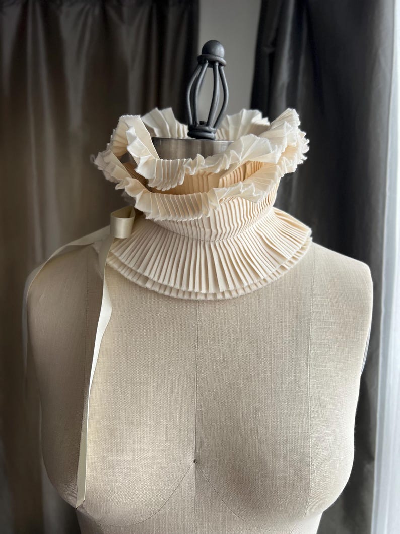 May include: A cream-coloured, pleated collar with a high neck and layered ruffles. A cream-coloured ribbon is attached to the side. The collar is displayed on a mannequin.