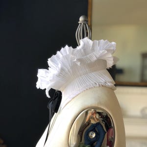 May include: A white, pleated ruffle collar with a black bow, displayed on a mannequin. The collar is made of a lightweight fabric. The mannequin is cream-coloured, and the background is dark.