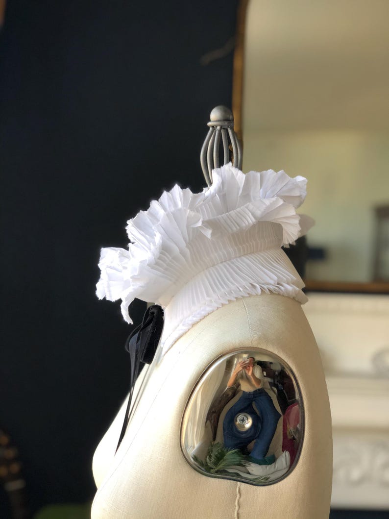 May include: A white, pleated ruffle collar with a black bow, displayed on a mannequin. The collar is made of a lightweight fabric. The mannequin is cream-coloured, and the background is dark.