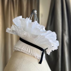 May include: A white, pleated collar with a black velvet ribbon, displayed on a mannequin. The collar has a ruffled, layered design, creating a voluminous effect. The background features neutral-toned curtains.