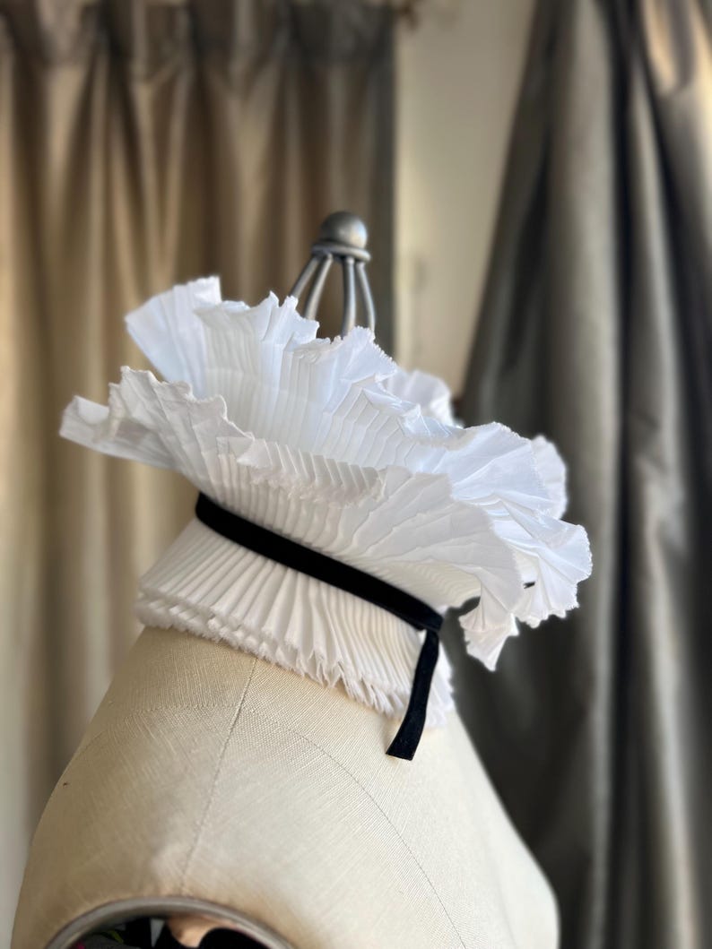 May include: A white, pleated collar with a black velvet ribbon, displayed on a mannequin. The collar has a ruffled, layered design, creating a voluminous effect. The background features neutral-toned curtains.