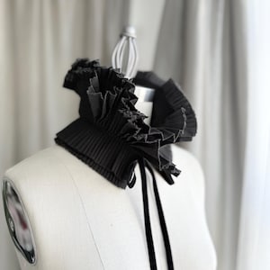 May include: A black pleated collar with a ruffled edge and a velvet tie, displayed on a mannequin. The collar has a structured design with a layered effect, creating a dramatic and elegant look. The background is a soft, neutral colour.