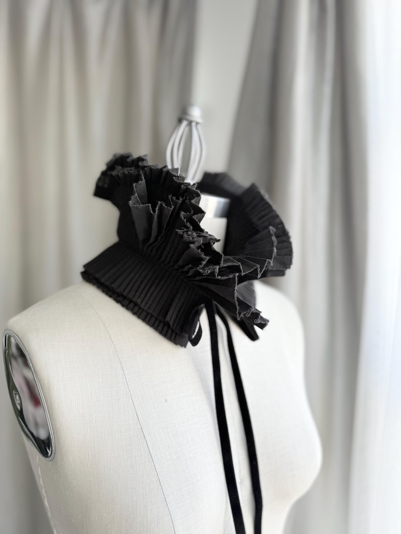 May include: A black pleated collar with a ruffled edge and a velvet tie, displayed on a mannequin. The collar has a structured design with a layered effect, creating a dramatic and elegant look. The background is a soft, neutral colour.