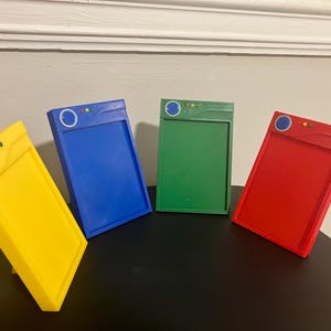 May include: Four colorful, rectangular objects in yellow, blue, green, and red. Each has a circular design and two small lights at the top. The objects are angled, with a black surface in the foreground and a white wall in the background.