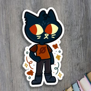 May include: A black cat character with red eyes and a brown shirt with a white symbol. The character is surrounded by autumn-themed decorations, including leaves and swirls.