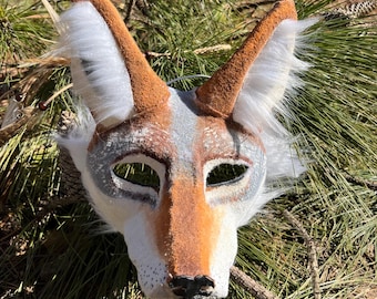 May include: A handcrafted coyote mask with a detailed design. The mask features a color palette of brown, white, and gray, with textured fur and pointed ears. The eye openings are oval-shaped. The mask is designed to be worn and is suitable for costume or decorative purposes.