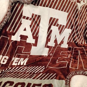 May include: A maroon and white fleece blanket with the letters "ATM" in white and the words "Gig 'Em" in white. The blanket has a striped pattern.