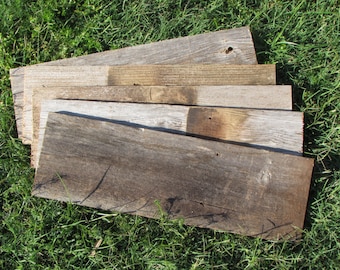 May include: A collection of reclaimed wood planks in varying shades of gray and brown, arranged on a bed of green grass. The weathered boards show natural grain and knots, suggesting a rustic or farmhouse aesthetic. The planks are of different lengths and widths.