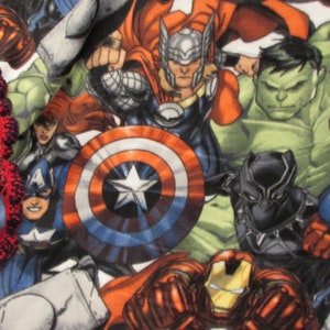 May include: A colorful fleece blanket featuring a variety of Marvel superheroes, including Captain America, Iron Man, Thor, Hulk, and Black Panther. The blanket is decorated with a red and black crocheted border.
