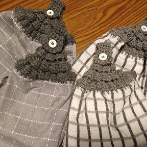 May include: Two gray and white kitchen towels with crocheted tops and button closures. One towel is solid gray with a white grid pattern, and the other is white with a gray grid pattern. The crocheted tops are dark gray.