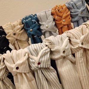 May include: A collection of hanging kitchen towels featuring a waffle-weave texture. Each towel is secured with a metal snap button closure, available in various colors including black, cream, teal, gray, orange, blue, and striped patterns.