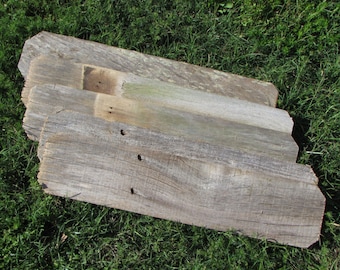 May include: Three weathered wooden planks stacked on top of each other, lying on a bed of green grass. The planks are gray and brown with visible knots and grain patterns.