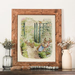 May include: A framed print featuring a watercolor illustration of rabbits in a garden setting. The artwork is set within a rustic, brown wooden frame. The scene depicts several rabbits near a garden gate and lush greenery.
