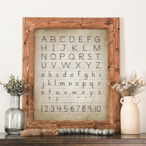 May include: A framed print with a rustic wooden frame displays the alphabet and numbers 1-10 in a classic, handwritten style. The background is a muted, vintage-inspired color. Decorative vases and beads are in the foreground.