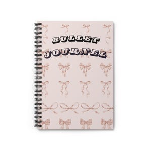 Puede incluir: Una libreta de espiral rosa con el texto "BULLET JOURNAL" en letras negras y negritas. La portada presenta un patrón repetitivo de lazos rosas.