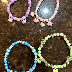 Op de afbeelding: Vier armbanden met kralen in verschillende kleuren. Eén is paars met kattenbedels, een andere is pastelkleurig met kattenbedels, een derde is blauw met vlinderbedels en de laatste is groen met kikkerbedels. De armbanden liggen op een donkerbruin oppervlak.