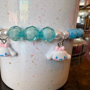 Op de afbeelding: Een armband met lichtblauwe, facetgeslepen kralen, witte parels en twee bedels. De bedels zijn wit met roze strikjes en blauwe details. De armband zit om een witte, getextureerde beker.