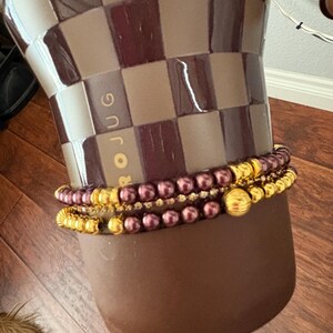 Op de afbeelding: Drie goud-burgundy armbanden zijn gestapeld op een bruine keramische vaas. De vaas heeft een dambordpatroon en het woord "JUG" erop gedrukt. De armbanden zijn een mix van kralen en gouden accenten.
