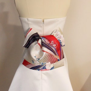 Può includere: Un abito da sposa bianco senza spalline con un grande fiocco colorato legato sul retro. Il fiocco è realizzato in tessuto fantasia con accenti rossi, bianchi, blu e dorati.