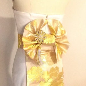 Può includere: Un manichino bianco senza spalline con una fascia e un fiocco floreale dorati. La fascia presenta un motivo floreale dorato ripetuto su sfondo crema. Un fiore e un nappa dorati decorativi accentuano il fiocco.