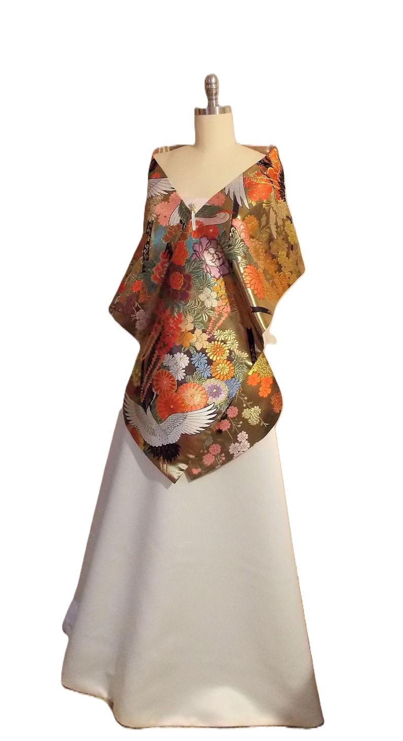 May include: A metallic gold shawl draped over a mannequin, featuring a vibrant pattern of orange, red, pink, and blue chrysanthemums, green foliage, and white cranes with black wingtips. The shawl is worn over a plain, floor-length white skirt.