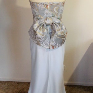Puede incluir: Un vestido azul claro sin tirantes con un corpi&ntilde;o ajustado y una falda fluida. El corpi&ntilde;o presenta un lazo decorativo hecho de una tela estampada con acentos dorados, blancos y marrones.