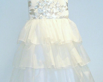 Puede incluir: Vestido blanco de tul en capas con un corpiño bordado en blanco y plata. El vestido es sin mangas y tiene una falda con volantes.