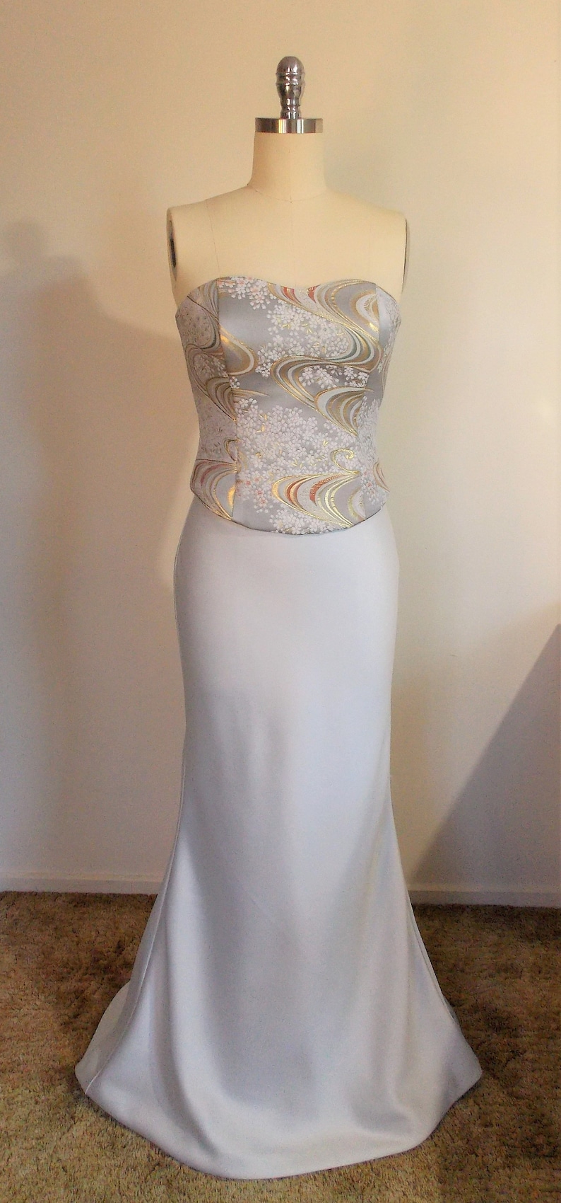 Puede incluir: Un vestido blanco sin tirantes con un corpi&ntilde;o ajustado y una falda fluida. El corpi&ntilde;o est&aacute; decorado con una tela estampada en plata y oro.