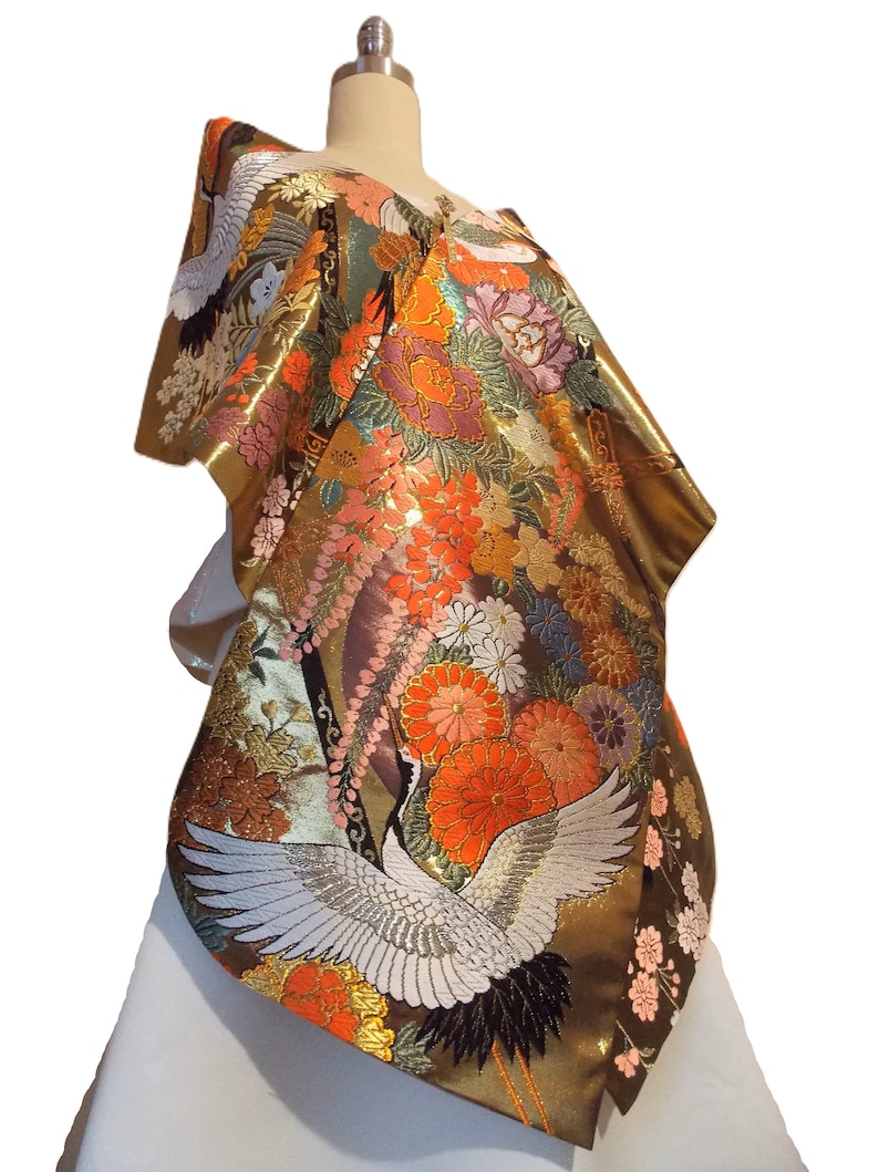 May include: A metallic gold kimono featuring intricate embroidery of white cranes with black-tipped wings, surrounded by vibrant orange, pink, and purple floral patterns including chrysanthemums and cherry blossoms on a shimmering fabric base.