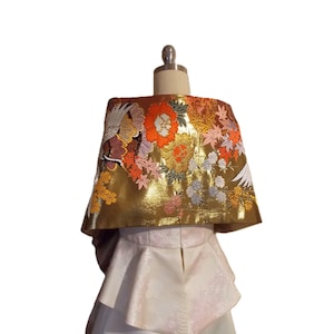 May include: A gold-colored wrap featuring intricate embroidery of white cranes, vibrant orange and red flowers, and maple leaves. The wrap is layered over a white, textured peplum-style garment with a subtle, repeating floral pattern.