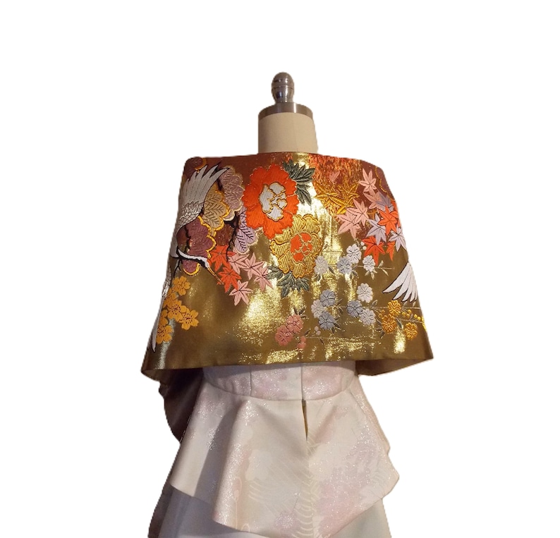 May include: A gold-colored wrap featuring intricate embroidery of white cranes, vibrant orange and red flowers, and maple leaves. The wrap is layered over a white, textured peplum-style garment with a subtle, repeating floral pattern.