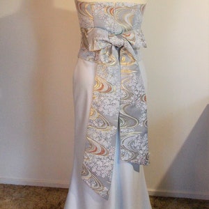 Puede incluir: Un vestido azul claro sin tirantes con una falda larga y fluida. El vestido tiene una faja estampada en blanco y dorado atada en un lazo en la espalda.
