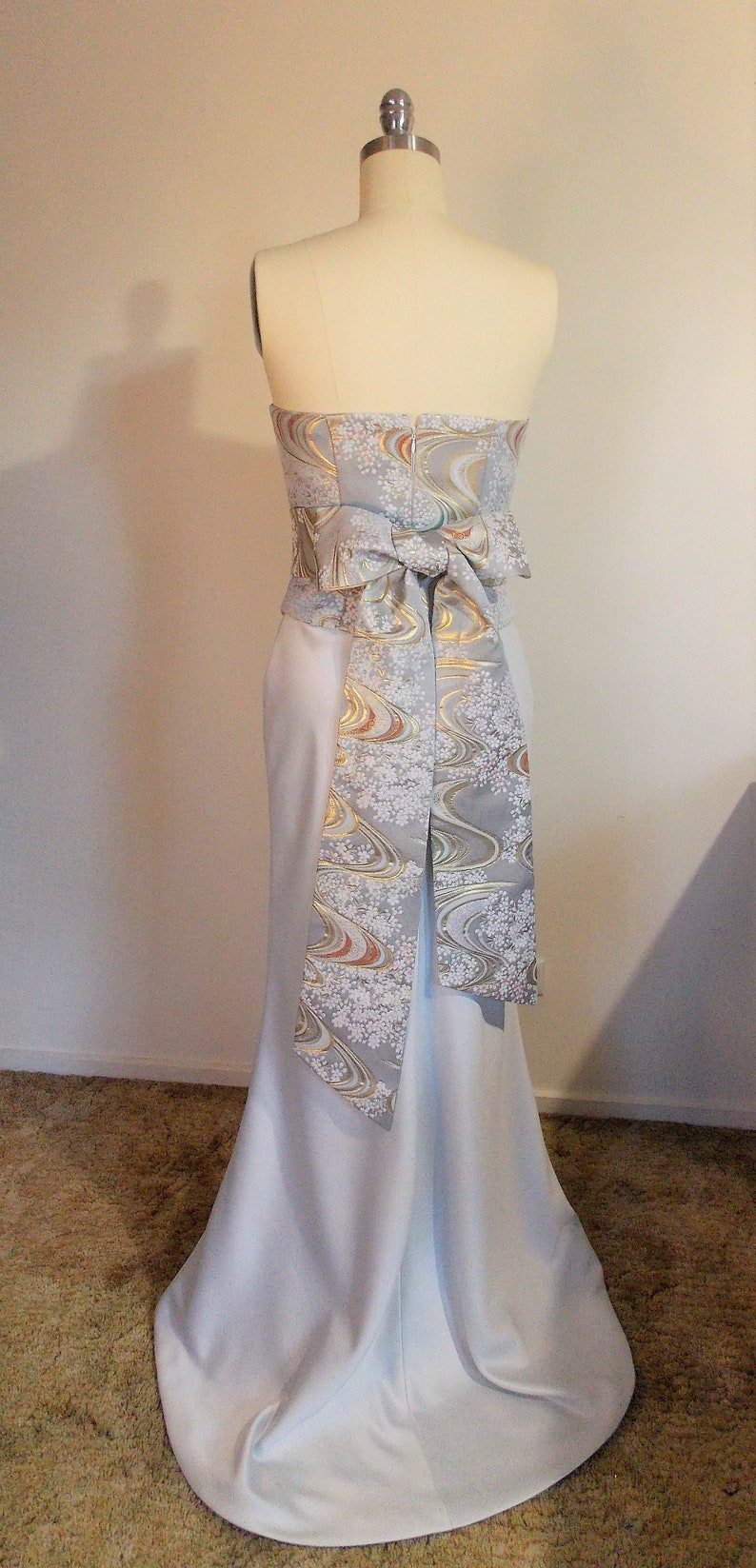 Puede incluir: Un vestido azul claro sin tirantes con una falda larga y fluida. El vestido tiene una faja estampada en blanco y dorado atada en un lazo en la espalda.