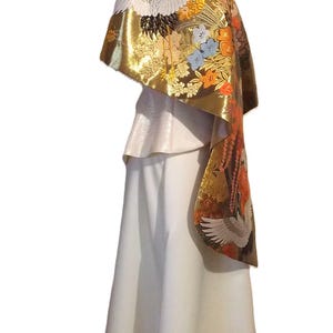 May include: A white floor-length skirt paired with a metallic gold shawl featuring intricate embroidery of white cranes, orange flowers, and blue blossoms. The shawl drapes over the shoulders, showcasing a detailed pattern of birds and floral motifs.
