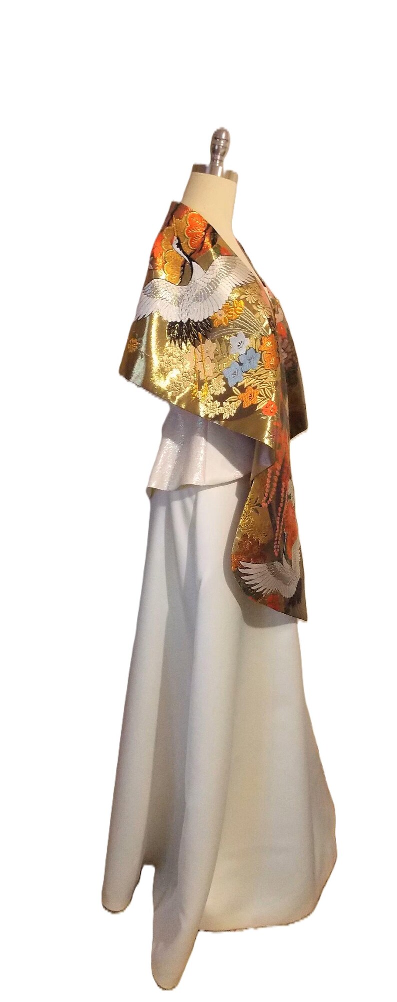 May include: A white floor-length skirt paired with a metallic gold shawl featuring intricate embroidery of white cranes, orange flowers, and blue blossoms. The shawl drapes over the shoulders, showcasing a detailed pattern of birds and floral motifs.