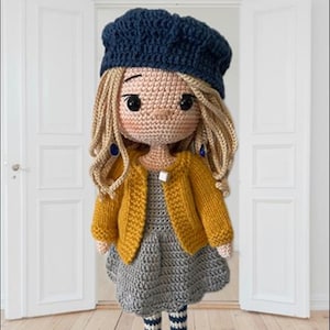 Könnte beinhalten: Eine gehäkelte Puppe mit einem blauen Barett, einem gelben Cardigan, einem grauen Kleid und gestreiften Socken. Die Puppe hat blonde Haare und braune Augen.