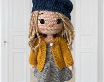 May include: A crocheted doll wearing a blue beret, yellow cardigan, gray dress, and striped socks. The doll has blonde hair and brown eyes.