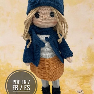 Puede incluir: Muñeca de ganchillo hecha a mano con gorro, chaqueta y botas azul marino. La muñeca tiene pelo rubio, una camisa a rayas y una falda naranja. La muñeca mide aproximadamente 30 cm de altura. La imagen incluye el texto "PDF EN / FR / ES".