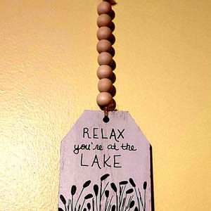 May include: Wooden lake-themed wall hanging with the text "RELAX you're at the LAKE". The sign is painted white with a blue base and black reeds. It is suspended by a string of wooden beads and a brown cord.